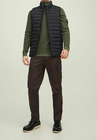 Mustavärinen toppaliivi vihreän neuleen päällä, yhdistettynä tummanruskeisiin cargo-housuihin ja mustiin kengiin, joissa on valkoinen pohja. Yksinkertainen muotoilu ja käytännölliset yksityiskohdat.