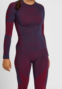 Nainen, joka pukee päälleen tiukan saumattoman pitkähihasen ykkösen ja legginsit tummanpunaisina ja laivastonsinisinä, joissa on geometrisia kuvioita.