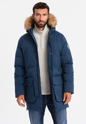 Chaqueta de plumas navy con capucha forrada de pelo, que presenta un cierre con cremallera en la parte delantera, dos grandes bolsillos con solapa y una silueta ajustada.
