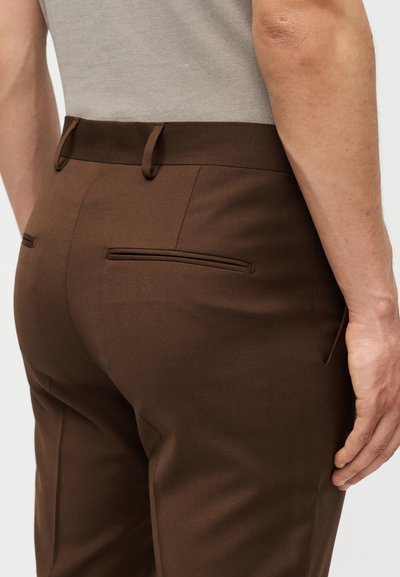 Κοντινό ενός ατόμου που φοράει καφέ παντελόνια με ραμμένες τσέπες στην πίσω πλευρά και μια μπεζ πουκαμίσα μέσα από το παντελόνι, με απλό φόντο.