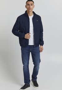 Marinblå vindjacka med dragkedja framtill, sidofickor och elastiska ärmslut, tillsammans med ljusa jeans och svarta sneakers.