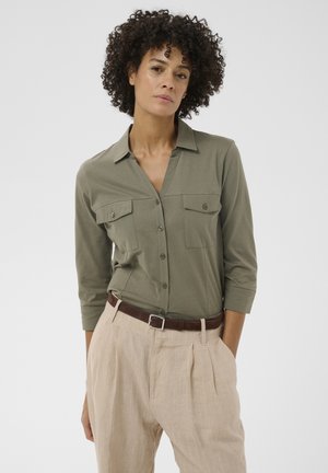 Woman with curly hair wearing an olive green button-up shirt and beige pleated pants with a brown belt, standing with one hand in pocket.