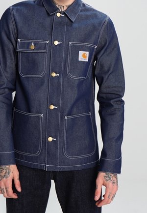 Homme portant une veste en jean Carhartt bleu foncé avec fermeture à boutons et quatre poches avant, montrant des tatouages sur ses deux mains et ses poignets.