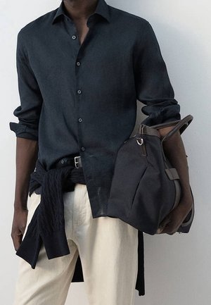 Persoon die een donker geknoopt overhemd en lichte broek draagt, met een donkere trui om de taille gebonden, die een donkere tas in één arm vasthoudt.