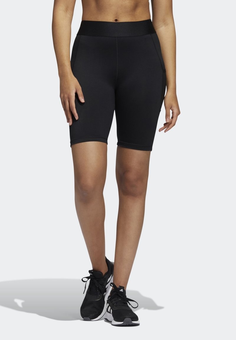 Shorts de sport noirs avec une taille haute, une texture lisse et une coupe ajustée, s'étendant jusqu'à mi-cuisse, associés à des chaussures de course noires.