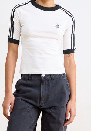 Osoba ubrana w białą koszulkę Adidas z krótkim rękawem, czarnymi paskami i logo, zestawioną z luźnymi ciemnoszarymi spodniami na białym tle.