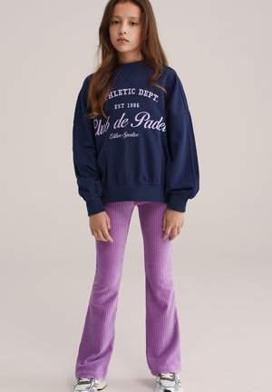 Sweat-shirt bleu marine avec texte blanc, associé à un pantalon évasé côtelé violet clair. Le modèle porte des baskets grises et blanches. Texture lisse.