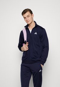 Chaqueta de chándal navy con cremallera y un alto collar, con el logo blanco de Adidas en el pecho, combinada con pantalones navy a juego y una bolsa de tirantes rosa.