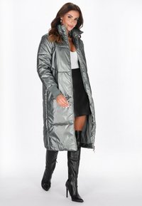 Polstret lang frakke i metallisk grå, med høj krave, lynlåslukning og ribbede manchetter. Sammen med en sort nederdel og knæ-høje støvler.