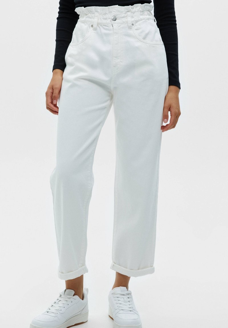 Weiße hochgeschnittene Jeans mit gerafftem Bund, zwei vorderen Taschen und umgeschlagenen Säumen, kombiniert mit weißen Sneakers. Sanfte Stoffstruktur.