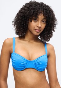 Γυναίκα με σγουρά μαλλιά που φοράει μπλε τοπ μπικίνι, κοιτάζοντας μπροστά με ουδέτερη έκφραση σε λευκό φόντο.