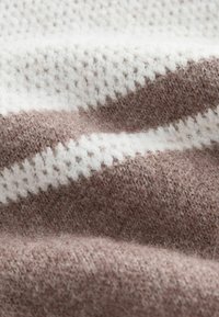 Strickdecke in Hellbraun und Weiß. Sie zeichnet sich durch ein strukturiertes Muster mit dicken und dünnen Streifen aus. Weiches, flauschiges Oberflächenmaterial.
