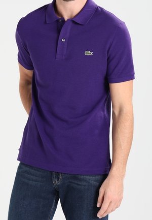 Uomo che indossa una polo a maniche corte viola con un piccolo logo di coccodrillo verde sul petto e jeans blu.