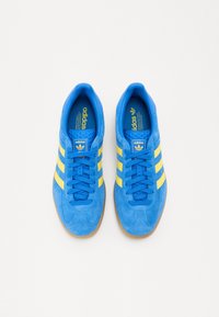 Blå ruskinds sneakers med tre gule striber, rund tå og gummisål. Branding på tungen og indersiden i gult. Snørebåndsdesign.