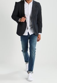 Dunkler Blazer über einem weißen Hemd mit kleinen blauen Punkten, kombiniert mit blauen Jeans und weißen Turnschuhen. Der Blazer hat einen Revers und aufgesetzte Taschen.
