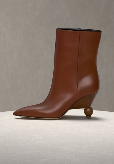 Καφέ δερμάτινο μποτάκι μέχρι τον αστράγαλο με μυτερή μύτη και μοναδικό στρογγυλεμένο γλυπτό τακούνι, εκτεθειμένο σε ανοιχτόχρωμη επιφάνεια πάνω σε ουδέτερο φόντο.