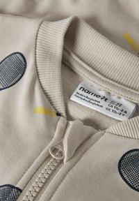 Beige Reißverschlusskleidungsstück mit einem gerippten Kragen. Verziert mit blauen runden Mustern und gelben Akzenten. Anhänger mit Pflegehinweisen und Größenangaben sichtbar.