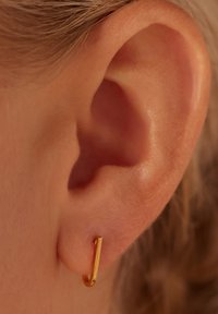Gouden creool oorbel, dun en gebogen, bevestigd aan een oorlel. Het ontwerp heeft een gladde oppervlakte met een lichte glans.