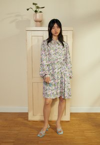 Vestido floral lilás claro com mangas bufantes longas e gola, apresentando padrões verdes e rosas. Usado com sapatos de salto azul com tiras.