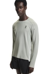 Maglietta sportiva a maniche lunghe in grigio chiaro, realizzata in tessuto traspirante. Presenta una scollatura tonda e un logo sul petto. Abbinata a pantaloncini neri.