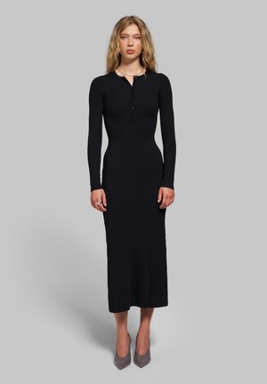Femme aux cheveux blonds ondulés portant une longue robe noire côtelée et des talons pointus gris sur un fond gris uni.