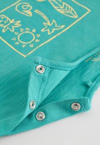 Turkoois stoffen kledingstuk met een zachte textuur, met gele geprinte ontwerpen van de zon, bladeren en fruit, en zilveren drukknoopsluitingen.