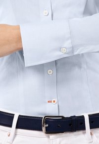 Camicia azzurro chiaro a maniche lunghe con bottoni bianchi, un asola cucita in arancione, abbinata a pantaloni bianchi e una cintura blu navy.