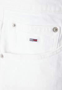 Hvite denimshorts med en liten brodert logo med røde og blå striper, metallknapper og en teksturert stoffoverflate.