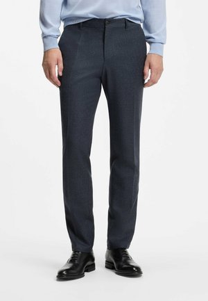 Hombre con pantalones entallados de color gris oscuro, zapatos de vestir negros y una camisa azul claro de manga larga, de pie contra un fondo blanco.
