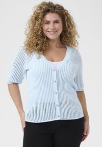 Cardigan tricoté bleu clair avec des manches courtes, texture en maille aérée, décolleté en V et grands boutons en perles sur le devant. Pantalons noirs portés en dessous.