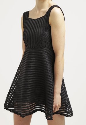 Kobieta ubrana w czarną sukienkę bez rękawów z poziomymi i ukośnymi koronkowymi wzorami w paski oraz rozkloszowaną spódnicą na jednolitym jasnym tle.