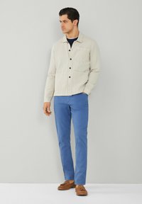 Chaqueta de botón en color beige claro con un bolsillo en el pecho, combinada con pantalones ajustados azules y mocasines marrones. Tela suave y diseño sencillo.