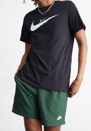 Personne portant un t-shirt noir Nike avec un double logo swoosh et un short Nike vert, debout avec une main dans la poche sur fond blanc.