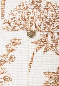 Tissu texturé crème avec un motif floral marron, bouton rond doré gravé "MOD" et détail matelassé subtil.