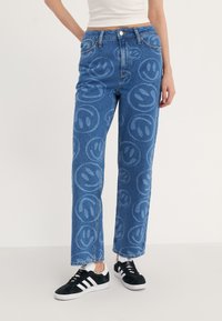 Jeans de mezclilla con un patrón repetido de caras sonrientes en azul claro sobre un fondo azul oscuro. Corte recto y cierre de botón.