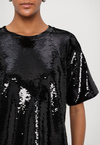 Black sequined short-sleeve top with a round neckline. The surface is textured with shiny, overlapping sequins creating a reflective effect.