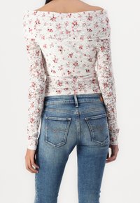 Mujer vistiendo una blusa de encaje blanca con volantes en los hombros, estampada con flores rojas, y jeans de mezclilla azul desgastados, vista desde la espalda.