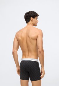 Muž s krátkými tmavými vlasy, bez trička, v černých vzorovaných boxerkách Calvin Klein, stojící proti světlému prostému pozadí, odvrácený zády.