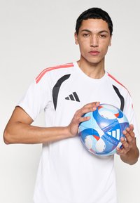 Λευκό αθλητικό πουκάμισο με μαύρες και κοραλλί λεπτομέρειες, κοντά μανίκια και στρογγυλή λαιμόκοψη· κρατώντας μια μπλε και κόκκινη ποδοσφαιρική μπάλα με σχέδιο.