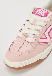 Lys rosa lædersneaker med hvide snørebånd og en gummirubber sål. Har lyserøde accenter og perforeringer på tåen for ventilation.