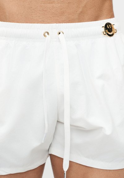 Λευκά σορτς με κορδόνι που φοριούνται στη μέση, με ένα χρυσό καρφίτσα με χαμογελαστό πρόσωπο στη δεξιά πλευρά της ζώνης.