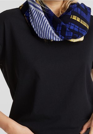 Personne portant une chemise noire à manches courtes et une écharpe infinie à motifs bleus, jaunes et blancs autour du cou.
