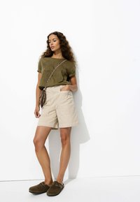 Chemise à manches courtes vert olive avec un design brodé, associée à un short cargo beige. Sabots en daim marron avec de la quincaillerie décorative.