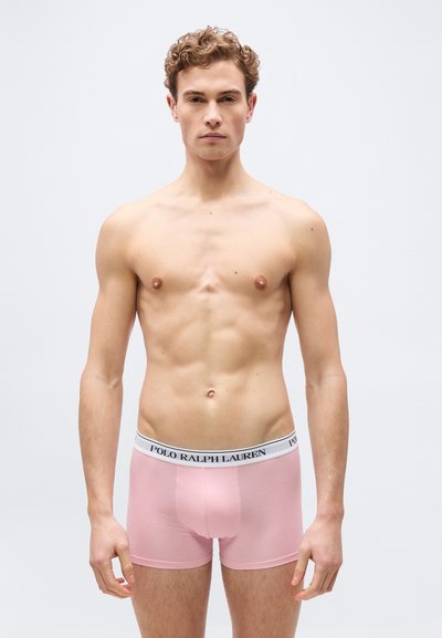 Άνδρας χωρίς πουκάμισο με σγουρά μαλλιά που φοράει εσώρουχα Polo Ralph Lauren σε ανοιχτό ροζ χρώμα, στέκεται μπροστά σε απλό φόντο.