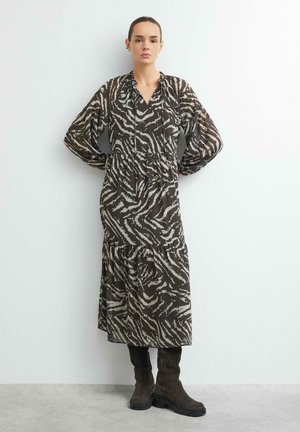 Frau trägt ein langärmeliges Midi-Kleid mit Zebramuster und dunklen Stiefeln, steht vor einer schlichten hellen Wand.