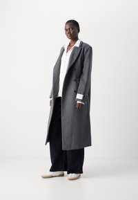 Palton gri din lână, cu design dublu-botonat, lung și supradimensionat, având butoane mari și o textură subtilă. Asortat cu o cămașă albă și pantaloni bleumarin.