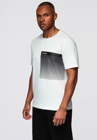 Bílá bavlněná trička s krátkým rukávem, které mají černý pruhoovaný design a kapsičku označenou "ombre." Jednoduchý a moderní střih.