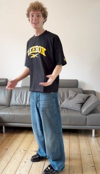 Juoda Reebok marškinėliai su geltonu logotipu, derinami su šviesiai mėlynomis plačiomis džinsų kelnėmis ir juodais batais. Išskiriama minkšta tekstūra ir laisvas kirpimas.