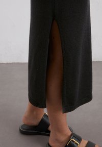 Schwarzes Strickkleid mit einem seitlichen Schlitz, ausgestattet mit gerippten Säumen. Kombiniert mit schwarzen Slide-Sandalen mit einem goldenen Schnallenakzent.