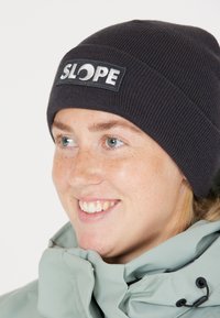 Svart ribbad mössa med en rektangulär lapp med ordet "SLOPE" i vitt. Mjuk tygkvalitet, sitter tajt runt huvudet.
